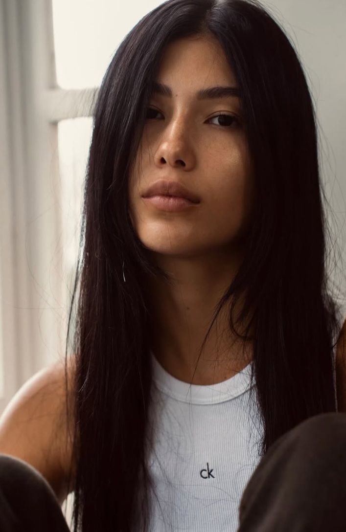 Woman with long black hair and freckles wearing a white tank top indoors