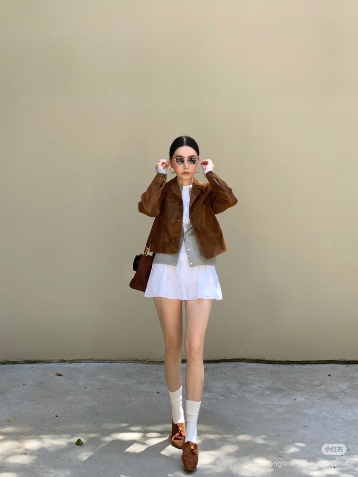 Woman in a brown jacket and white skirt adjusting her sunglasses outdoors