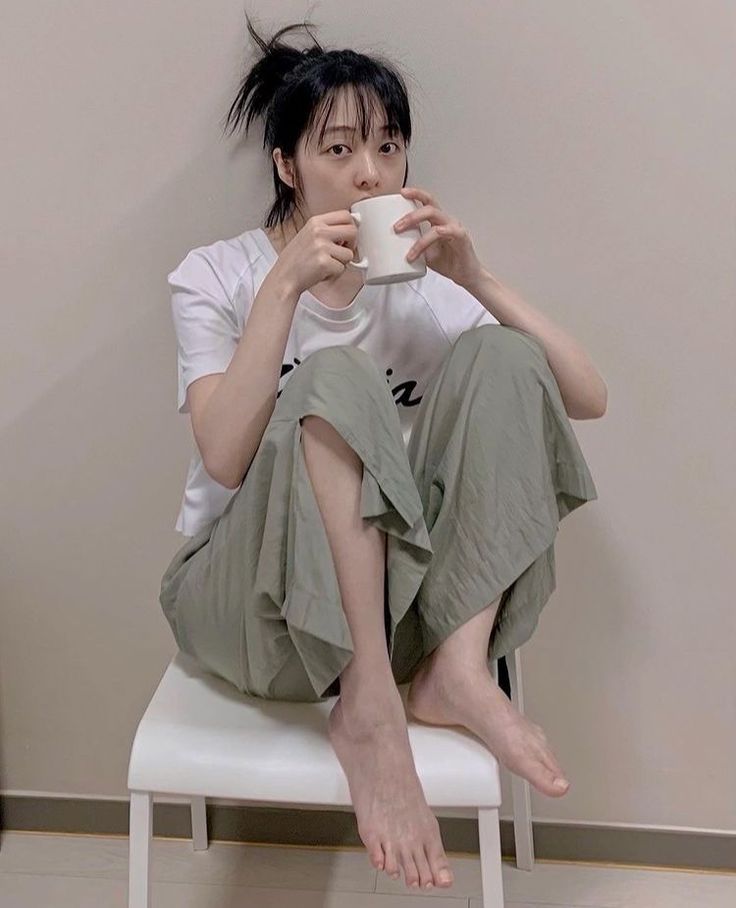 Woman with black hair in a messy bun, sitting on a chair and drinking from a mug