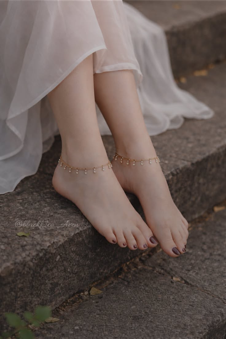 Bare feet with anklets and dark nail polish, wearing a white dress outdoors