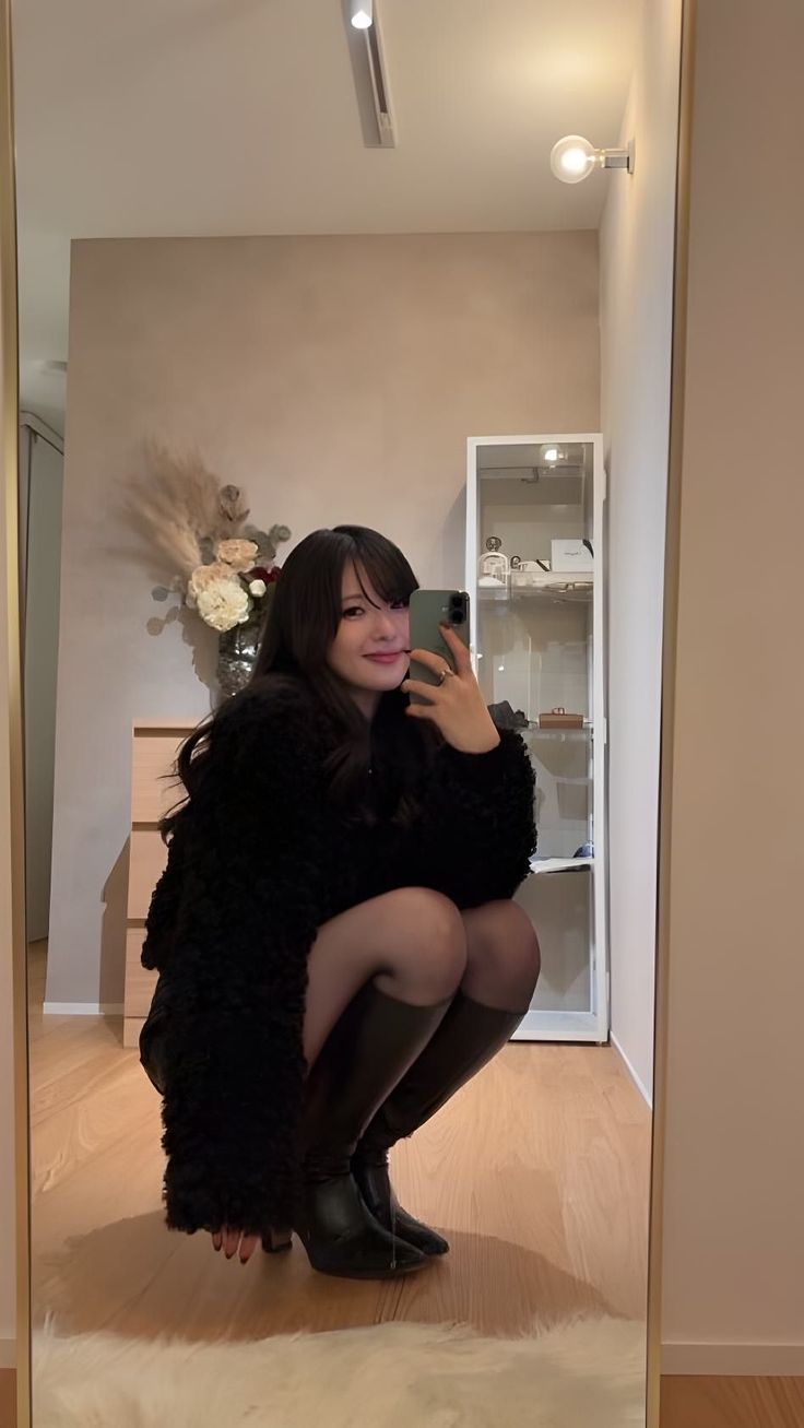 Woman in a black jacket and boots taking a mirror selfie indoors