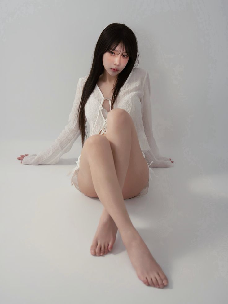 Woman with long dark hair wearing a white top and shorts, sitting on a white floor