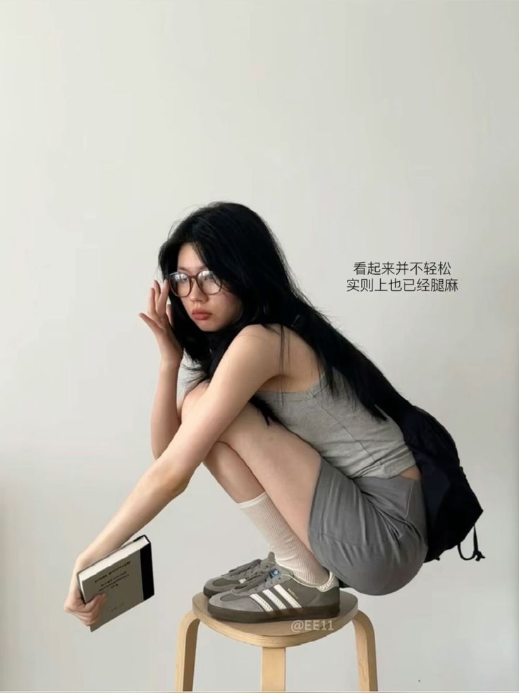 Woman with long black hair and glasses squatting on a stool, holding a book
