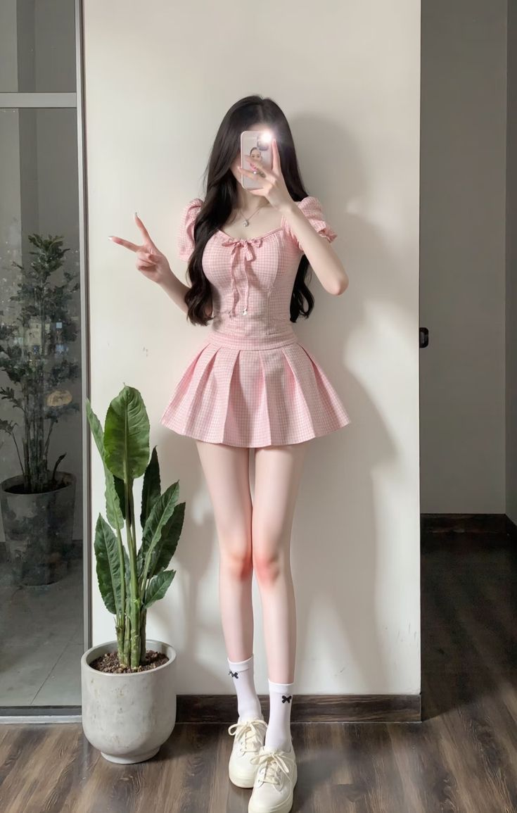 Woman in a pink gingham top and pleated skirt taking a selfie indoors