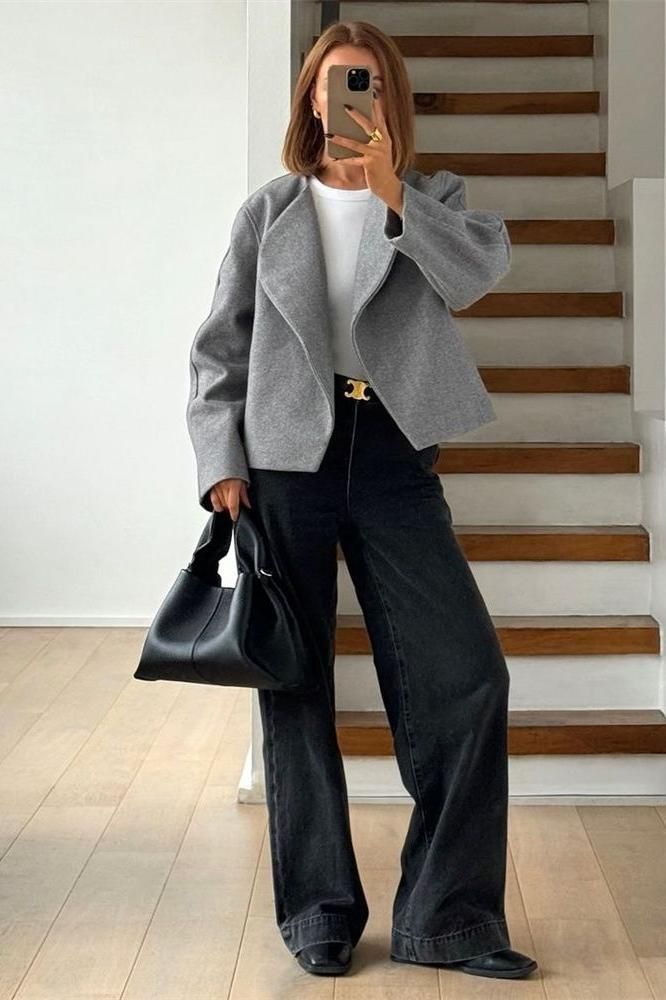 Woman in a gray jacket and wide leg pants holding a black handbag indoors