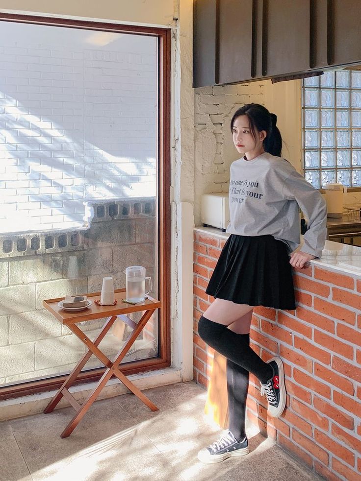 Woman in a grey long sleeve shirt and black pleated skirt leaning against a brick wall