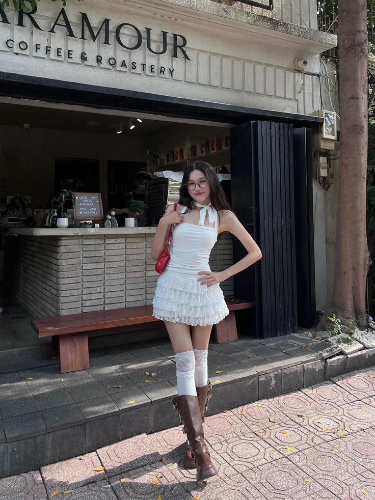Woman in a white dress, brown boots, and glasses standing outside a coffee shop