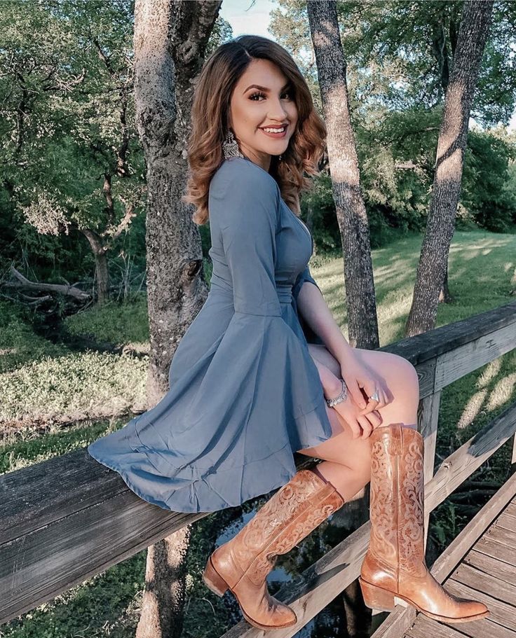 Woman in a blue dress and cowboy boots sitting on a wooden fence outdoors