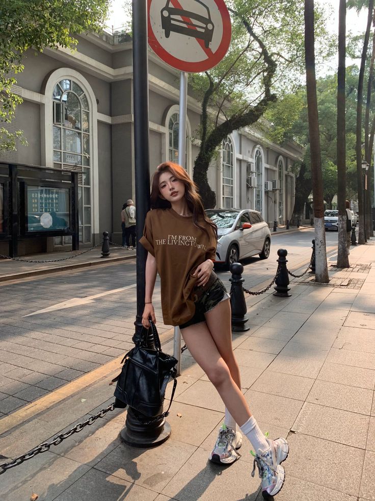 Woman in a brown t-shirt and denim shorts leaning on a lamppost on a street