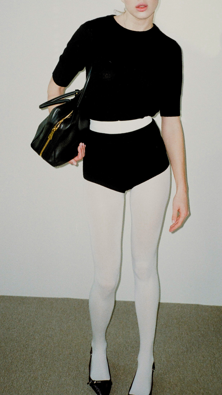 Woman in a black top, black shorts, and white tights holding a black handbag