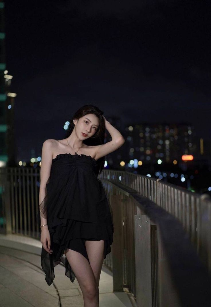 Woman in a black dress posing on a bridge at night with city lights in background