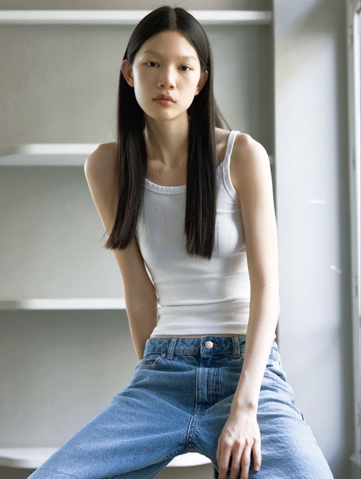 Woman with long black hair wearing a white tank top and blue jeans indoors