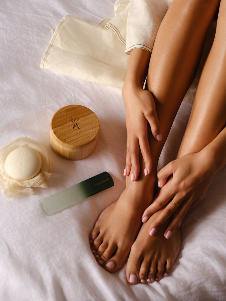 Person's feet and hands with pink nail polish on white bedding with beauty products
