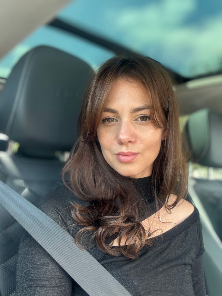 Woman with long brown hair in a car, wearing a black top and seatbelt