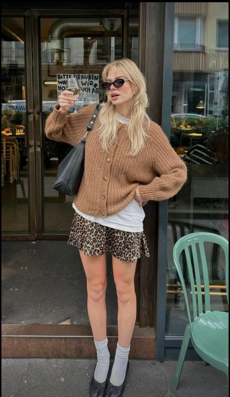 Blonde woman in a brown cardigan and leopard print skirt holding a drink outdoors