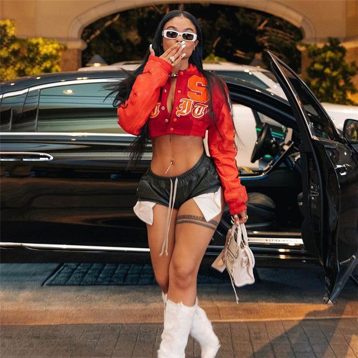 Woman in a red cropped jacket and short shorts next to a car
