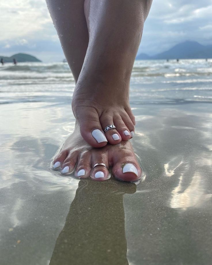 Feet with white nail polish and toe rings standing in shallow ocean water on a beach