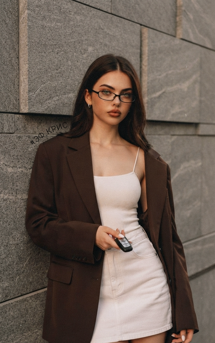 Woman with long hair and glasses wearing a brown blazer and white skirt outdoors