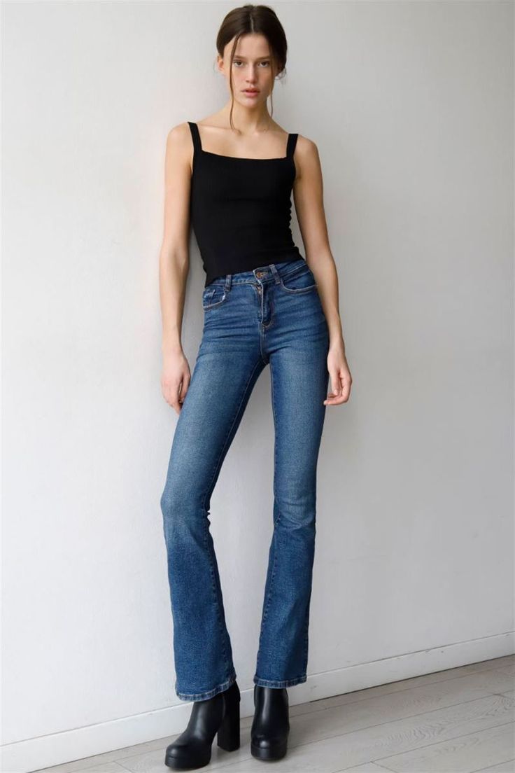 Woman in a black tank top and blue jeans leaning against a white wall