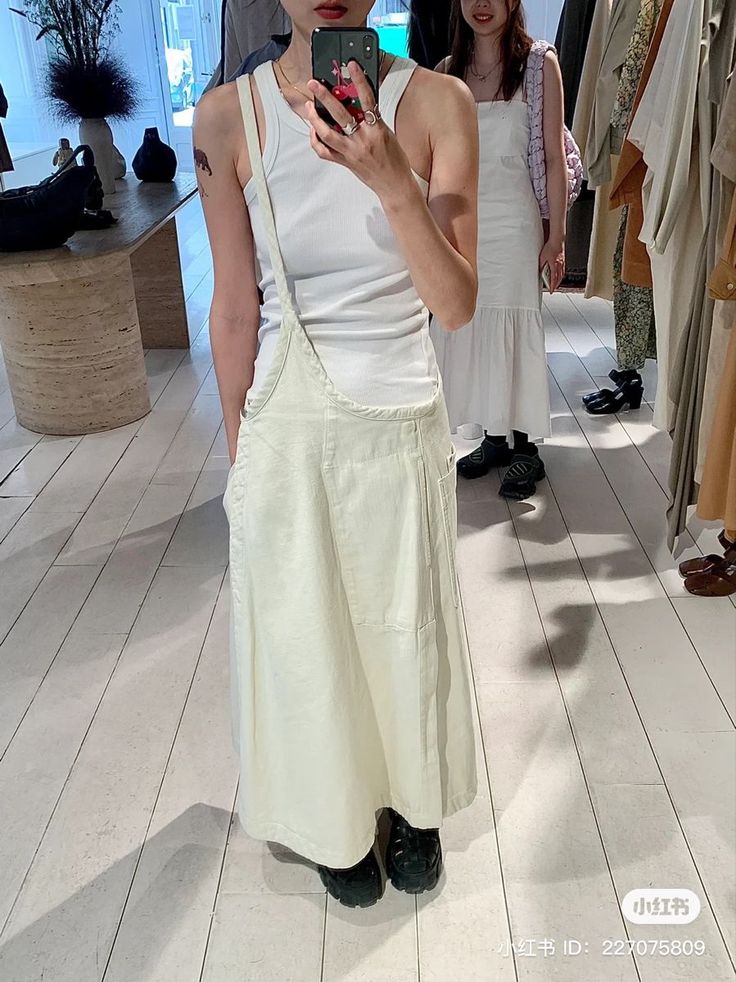 Person in a white tank top and long skirt overalls taking a mirror selfie in a store