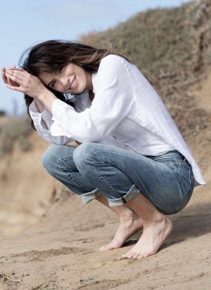 Woman in a white shirt and jeans squatting barefoot on sand, smiling