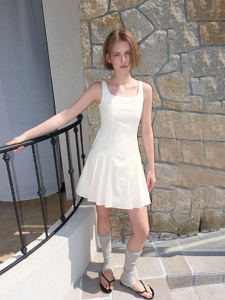 Woman in a white dress and light socks standing outdoors by a stone wall
