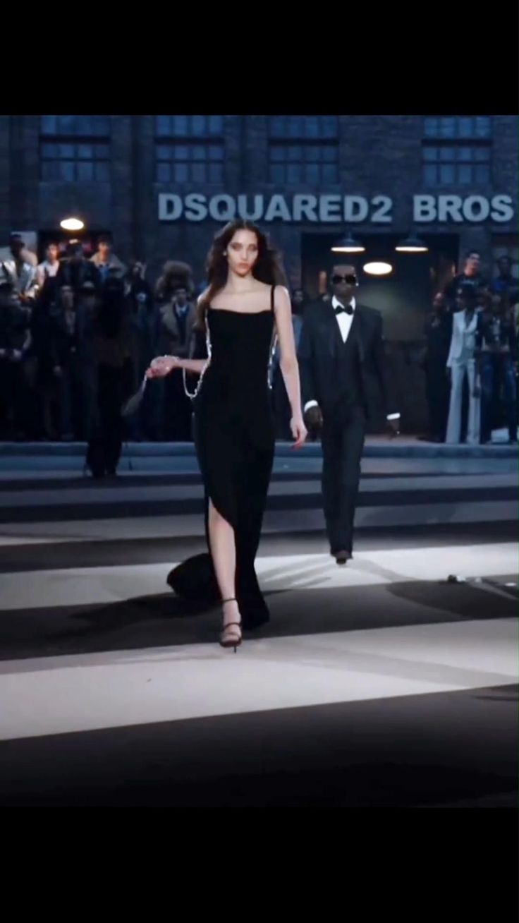 Woman in a black dress walking on a runway with a man in a suit behind her