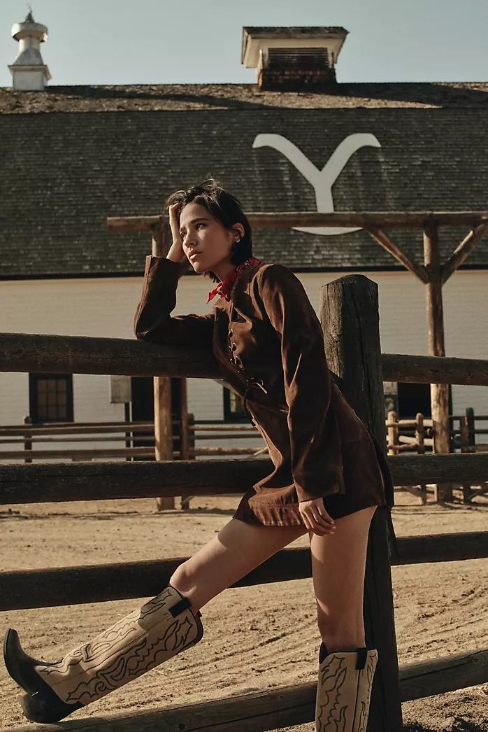 Woman in a brown jacket and cowboy boots leaning on a wooden fence at a ranch