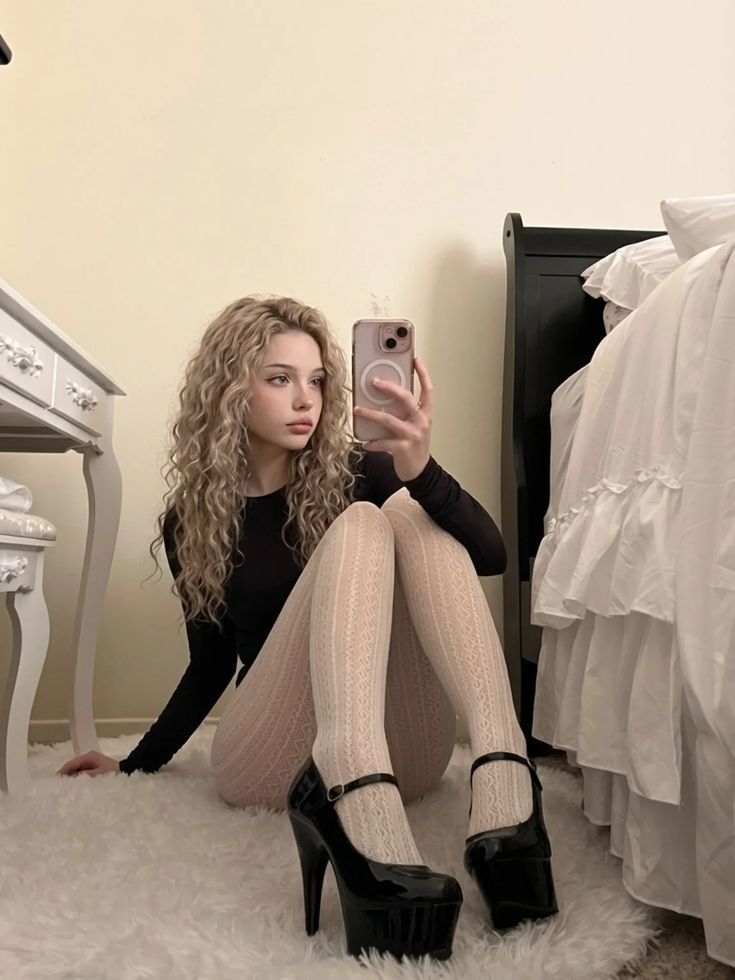 Woman with curly blonde hair taking a mirror selfie in a bedroom