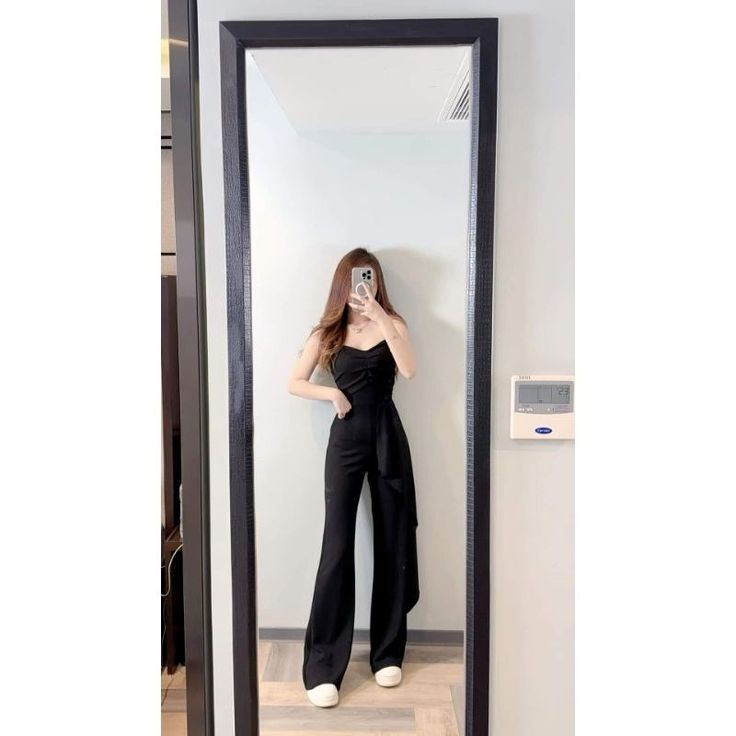Woman in a black jumpsuit taking a mirror selfie indoors