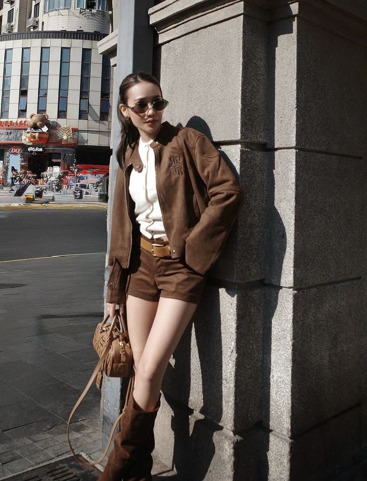 Woman in a brown jacket and shorts leaning against a wall on a sunny street