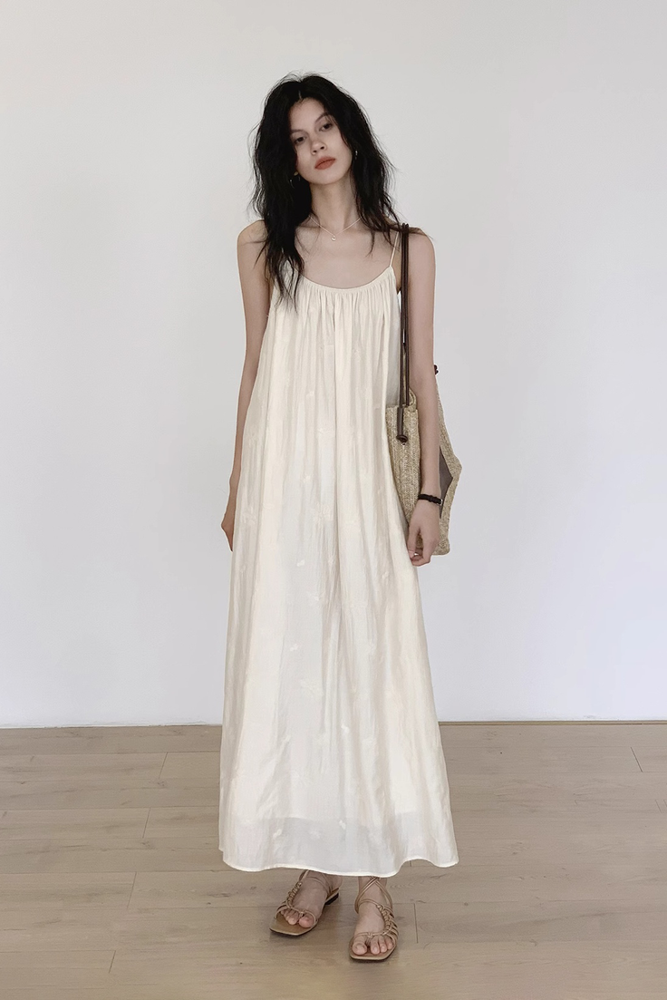 Woman in a long cream dress and sandals holding a straw bag indoors