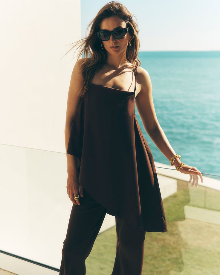 Woman in sunglasses and a brown outfit standing on a balcony overlooking the ocean