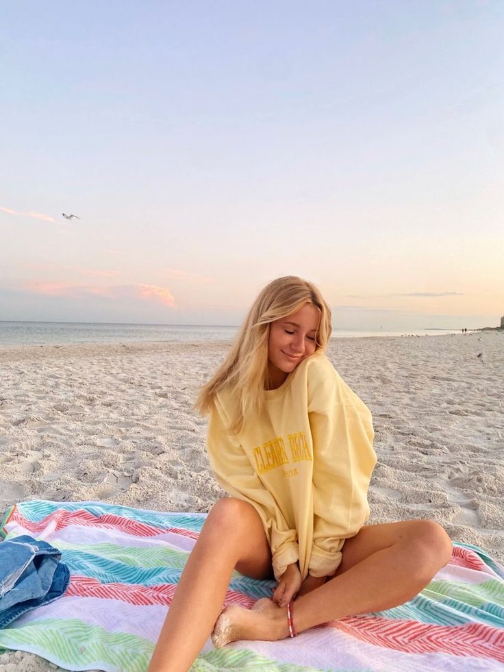 Blonde woman in an oversized yellow sweatshirt sitting on a beach at sunset