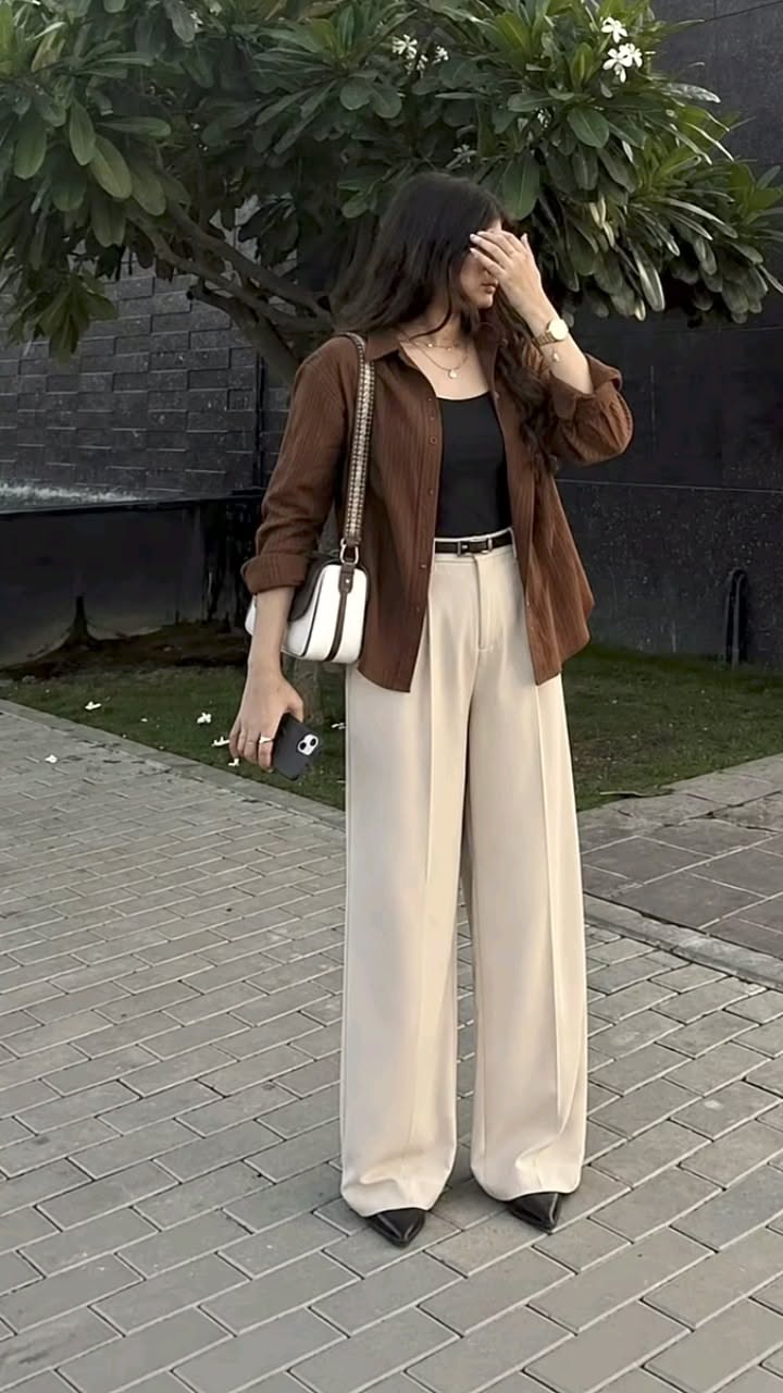 Woman in a brown shirt and wide leg pants holding a phone outdoors