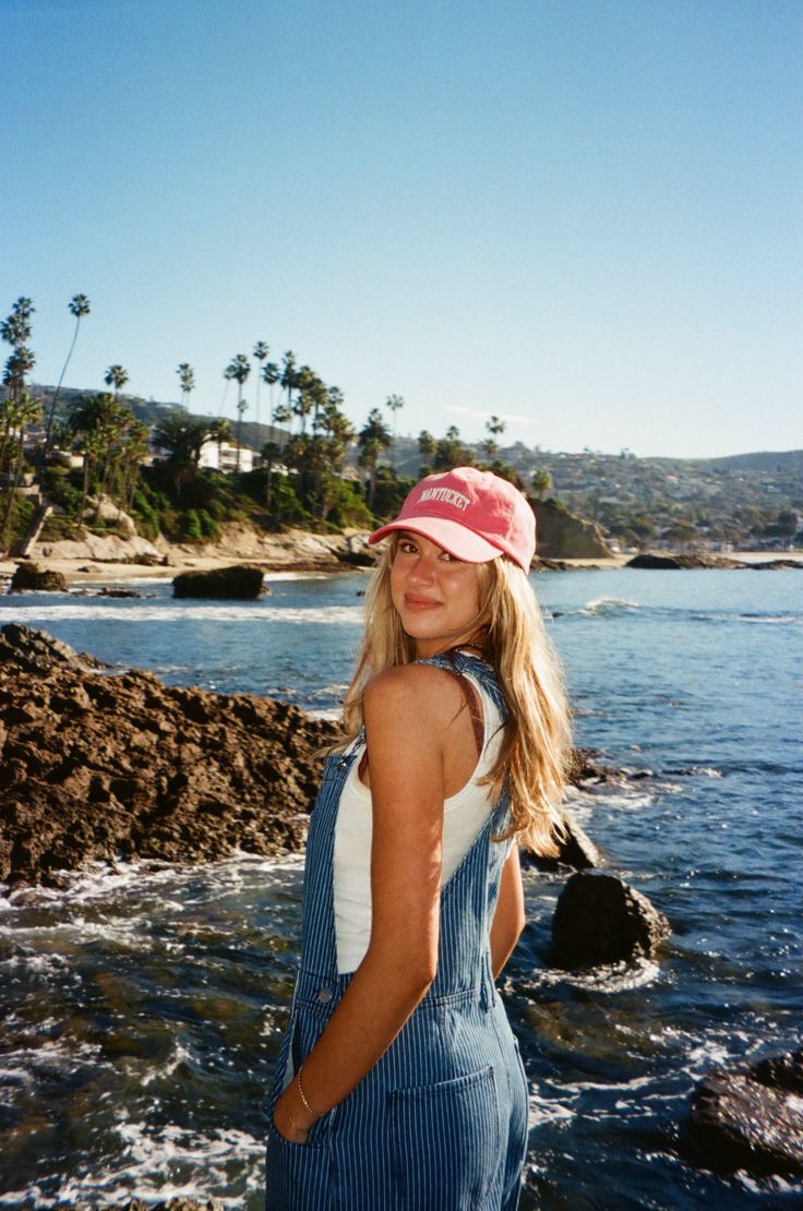 Young woman in denim overalls and a pink cap at a rocky beach with palm trees