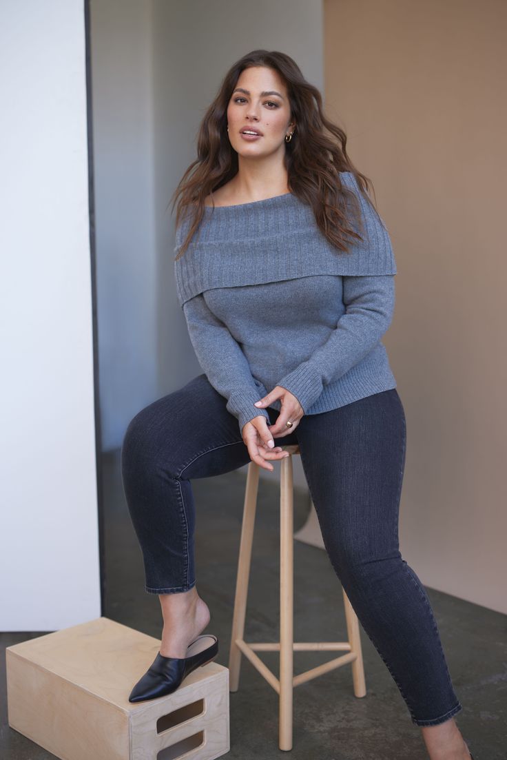 Woman with long hair wearing a gray off-shoulder sweater and dark jeans, sitting on a stool