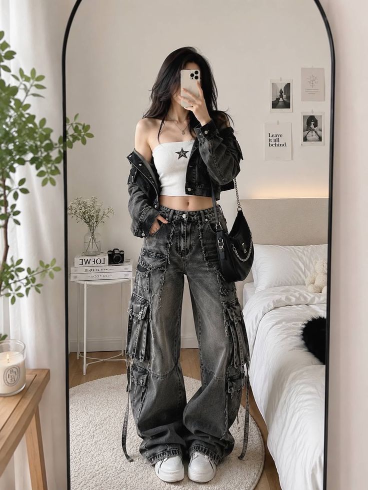 Person in a crop top, denim jacket, and cargo pants taking a mirror selfie in a bedroom