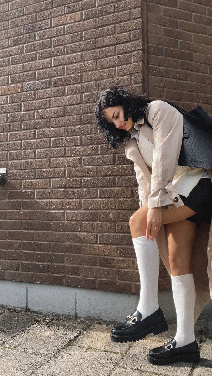 Woman in a trench coat, black shorts, and white socks leaning against a brick wall