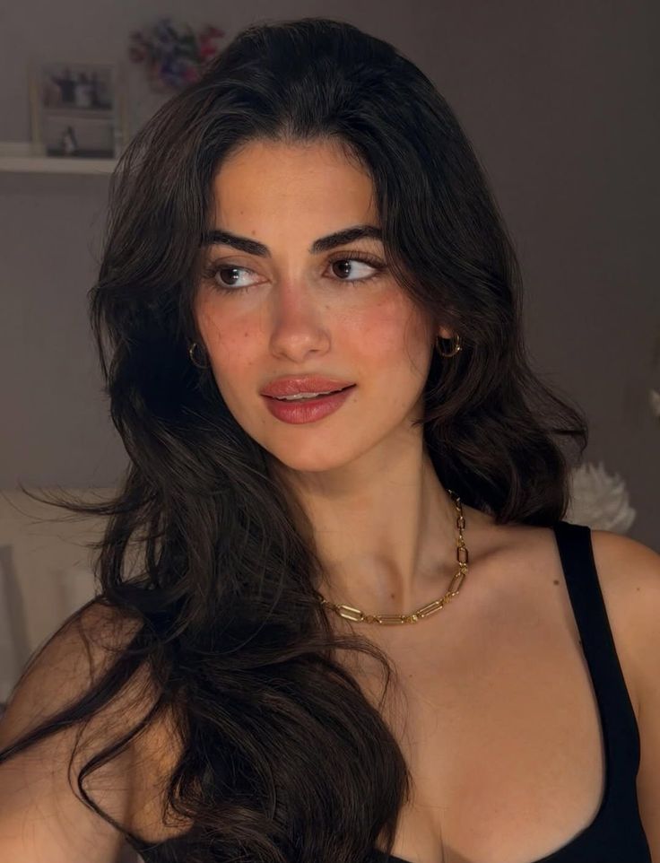 Woman with long dark hair wearing a gold necklace and black top indoors