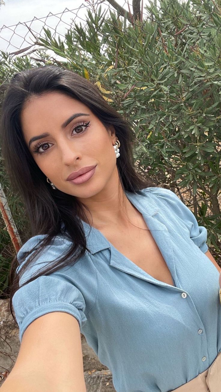 Woman with long dark hair and pearl earrings wearing a blue shirt outdoors