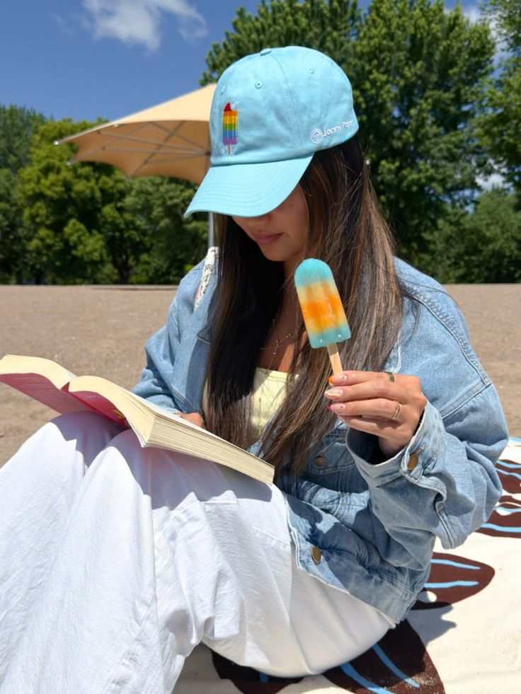 Woman in a denim jacket and baseball cap reading a book and holding a popsicle