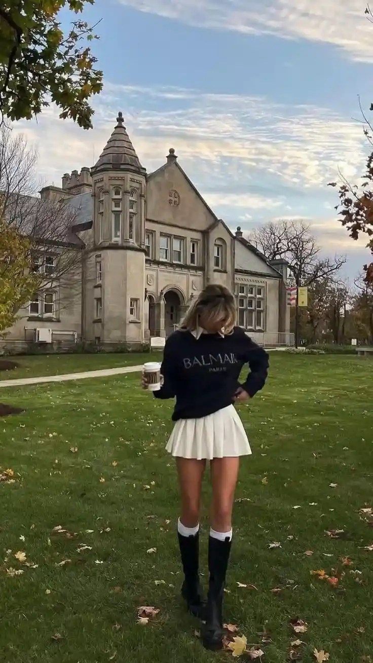Woman in a pleated skirt and long boots holding a cup in front of a historic building