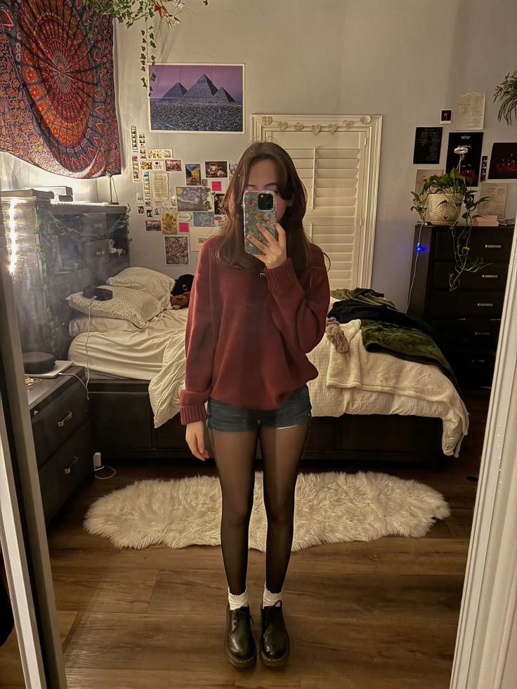 Person in a red sweater and denim shorts taking a mirror selfie in a bedroom