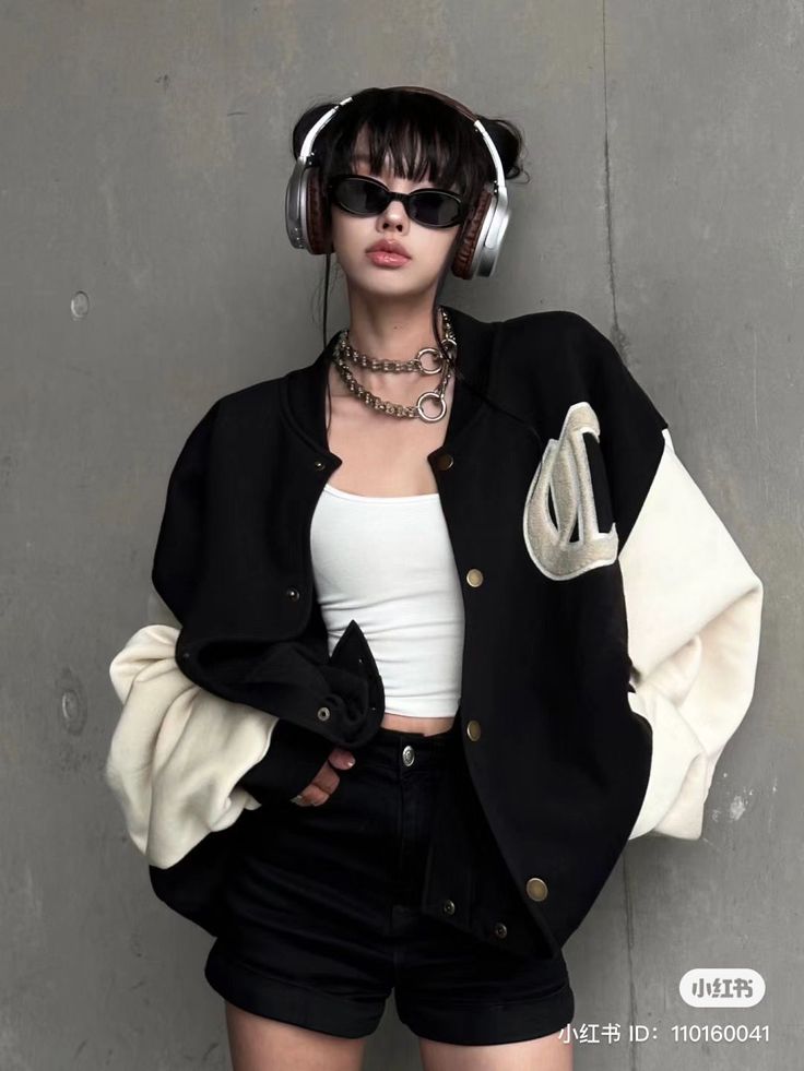 Person in a black and white varsity jacket, headphones, and sunglasses against a gray wall