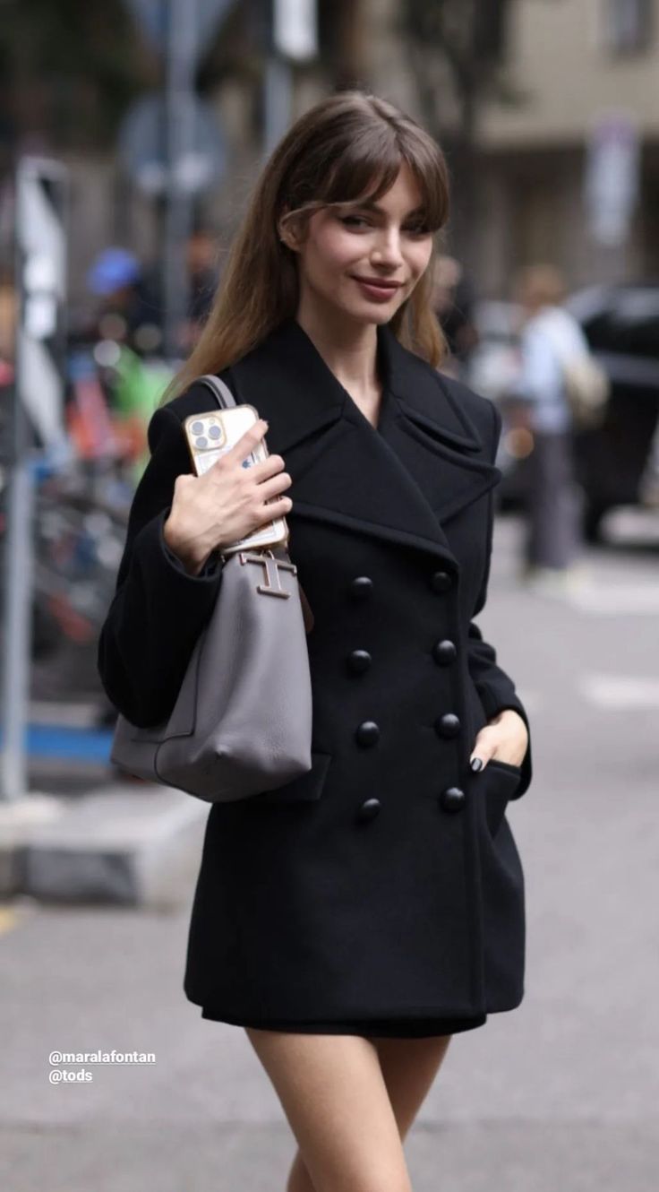 Woman in a black coat holding a grey handbag and phone on a street