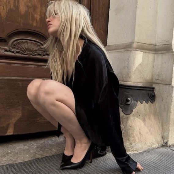 Woman with long blonde hair in a black dress and high heels squatting outdoors