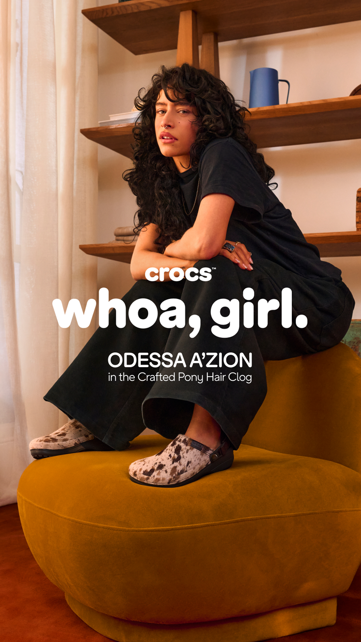 Woman with long curly hair wearing a black t-shirt and cow print clogs, sitting indoors