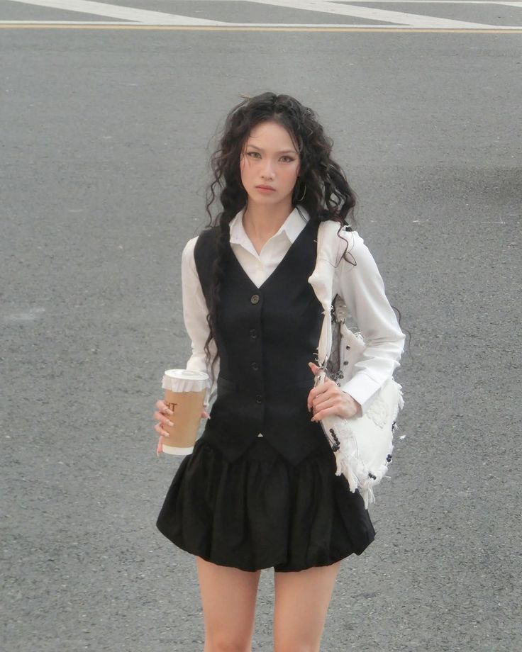 Woman with long curly hair in a black vest and skirt holding coffee on a street