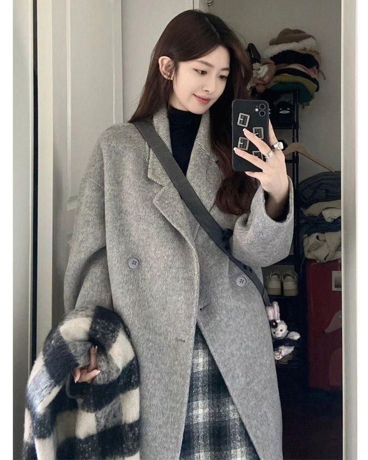 Woman in a gray coat and plaid skirt taking a mirror selfie indoors